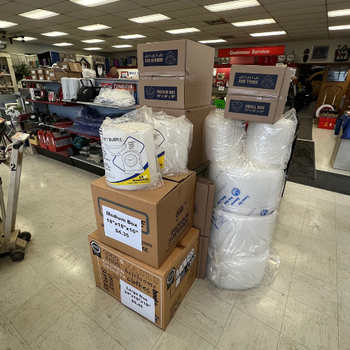 A store interior with packing supplies, including stacked boxes, rolls of bubble wrap, and packing paper. Shelves display various household items.