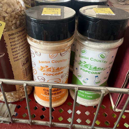 Two bottles of Amish Country popcorn seasoning are displayed on a shelf. One is cheddar cheese flavored, the other is butter flavor.