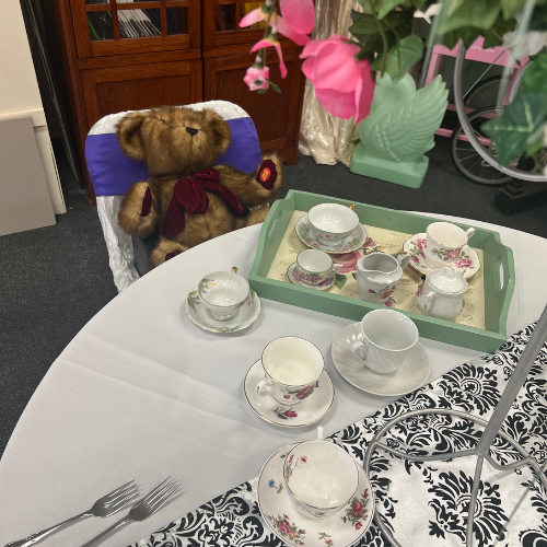 A plush teddy bear sits at a table set for tea with floral-patterned china and a decorative tablecloth. Pink flowers and a peaceful atmosphere enhance the charm.
