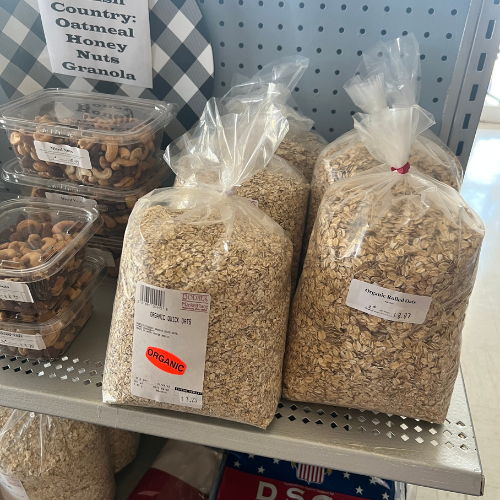 Plastic bags filled with organic oats and pretzels sit on a store shelf beside containers of mixed nuts. Labels display pricing and product information.