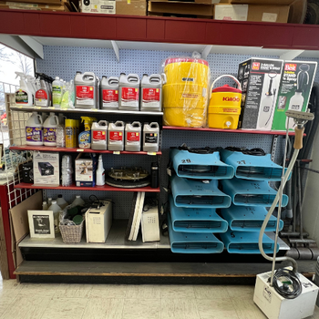 A store shelf with cleaning supplies, including jugs of chemicals, spray bottles, a yellow Igloo container, and blue air movers. The setting is organized and utilitarian.