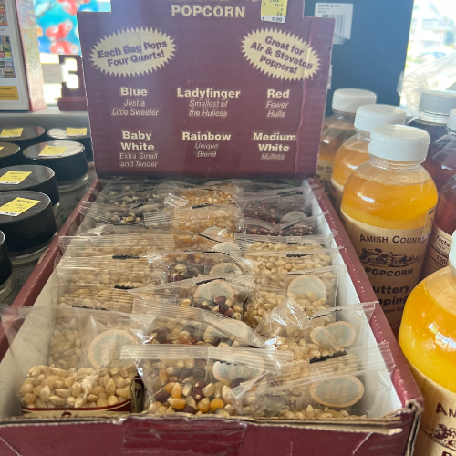 Box of assorted popcorn kernels, each variety labeled: Blue, Ladyfinger, Red, Baby White, Rainbow, Medium White. Set beside bottles of Amish Country popcorn seasoning.