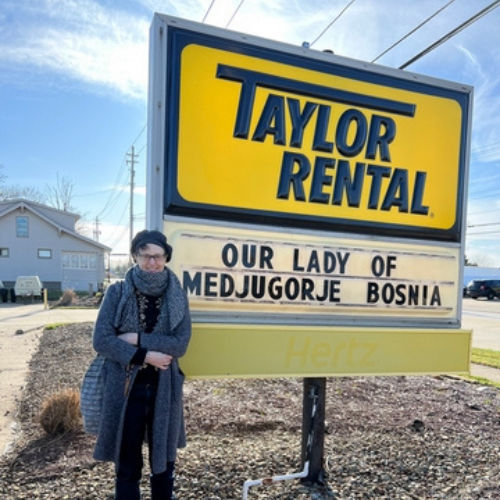 A person in a cozy outfit stands smiling next to a large sign for 
