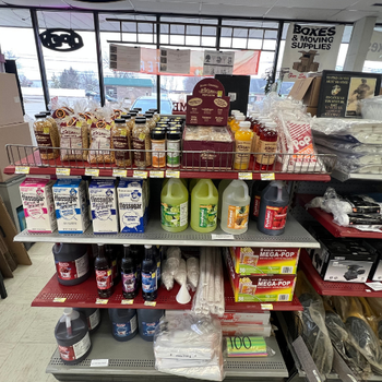 Store shelf with various popcorn supplies. Top shelf has popcorn kernels and flavorings. Middle shelf has flossugar containers and syrup bottles. Bottom shelf contains cups, bags, and a popcorn machine box. Brightly lit setting.
