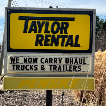 Sign for Taylor Rental features a bright yellow background, announcing, "We now carry U-Haul trucks & trailers." The mood is informative and promotional.